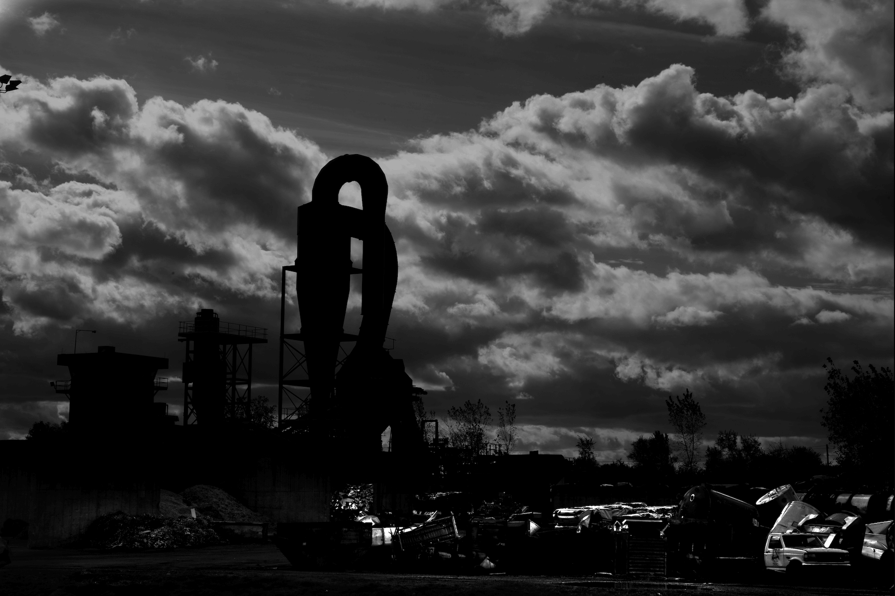 a silhouette of a massive metal re-cycling machine with a pile of dead cars in the foreground