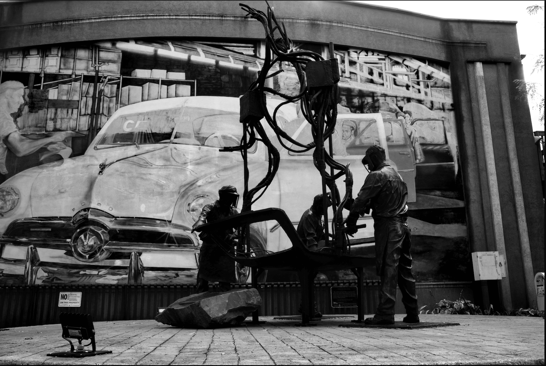 a silhouette of three statue of three automotive workers with a mural of a abstract car in the background